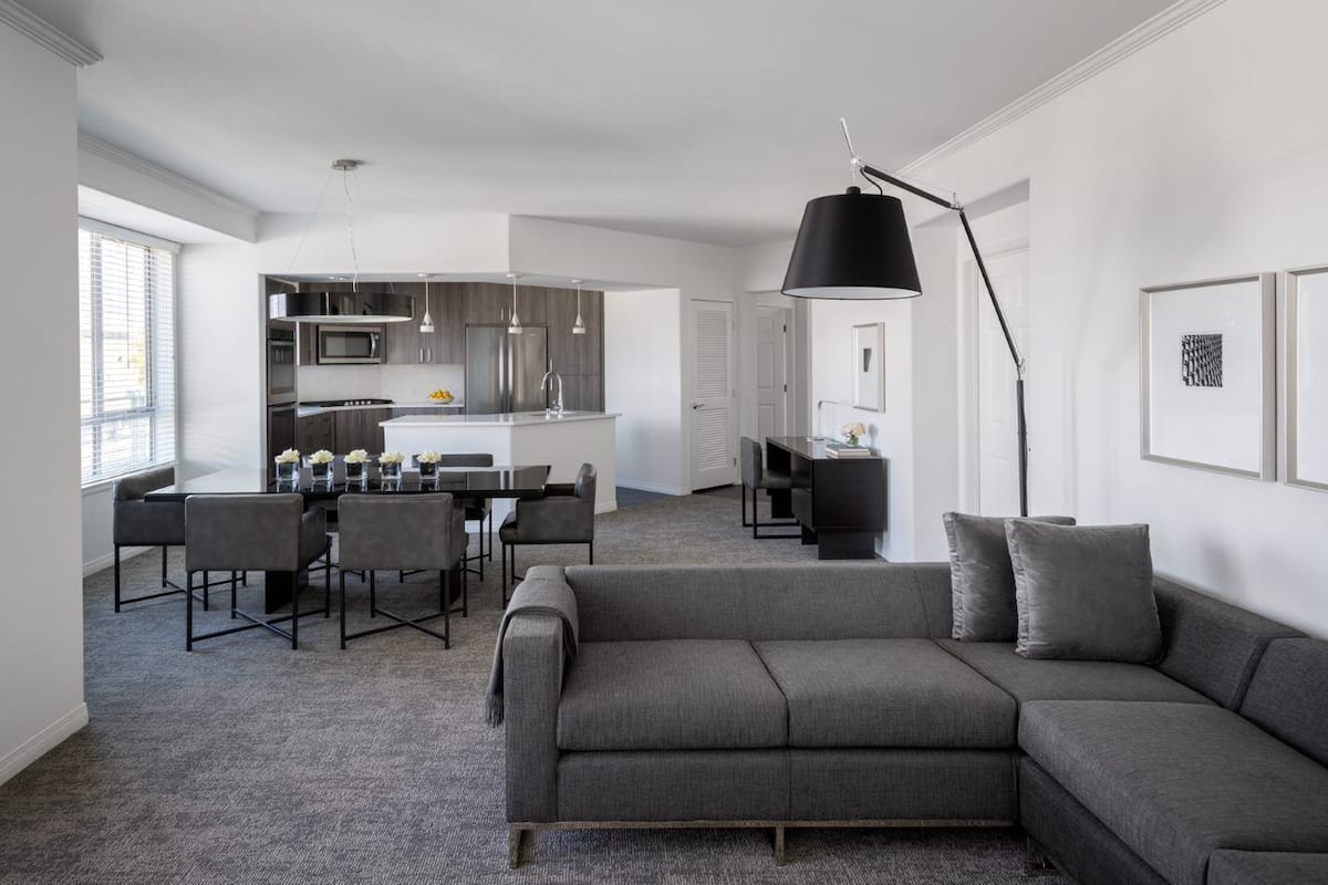 A modern living room with a grey sofa, dining area with chairs, a kitchen with stainless steel appliances, and a large black floor lamp.