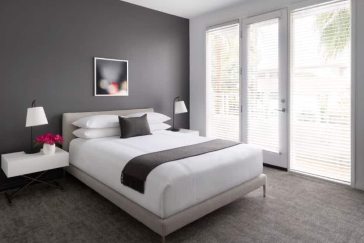 A modern bedroom with a large bed, two bedside tables, a nightstand, a wall-mounted lamp, and a window with blinds.