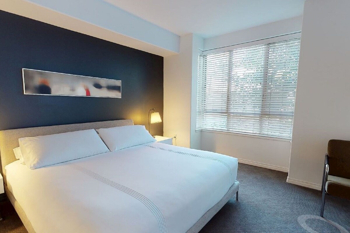 A hotel room with a large bed, two bedside tables with lamps, a window with blinds, and a chair.