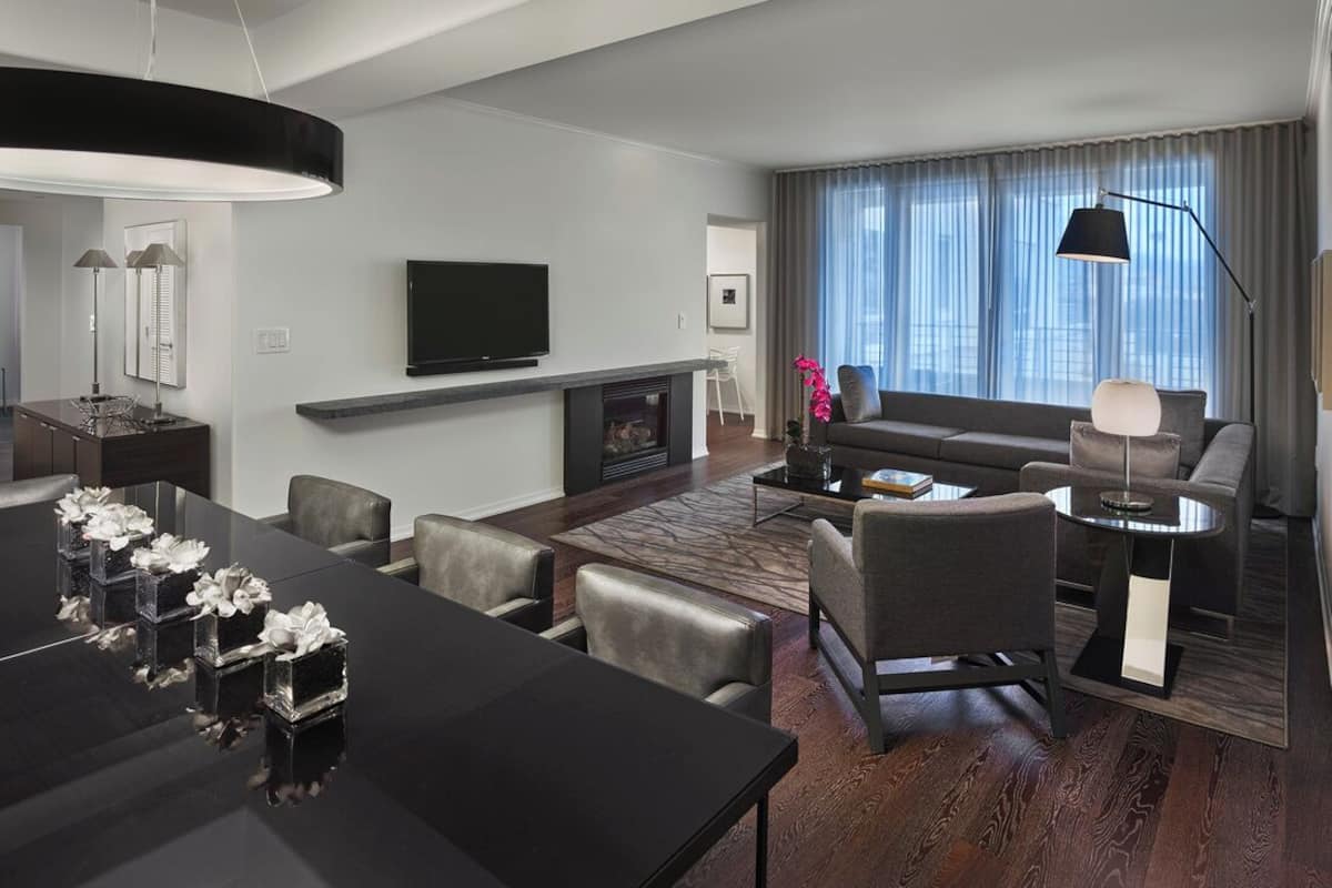 A modern hotel room with a dining area, a sofa set, a flat-screen TV, and a large window with curtains.