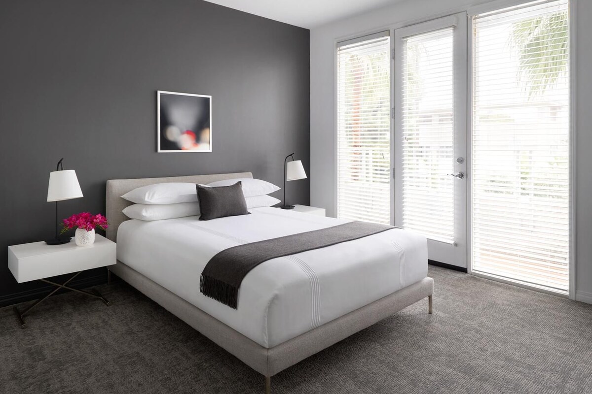 A modern bedroom with a large bed, two bedside tables, a nightstand, a picture on the wall, and a view through the window.