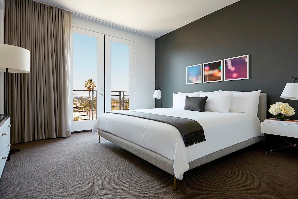 A hotel room with a large bed, a desk with a lamp, a view of the outdoors, and artwork on the wall.