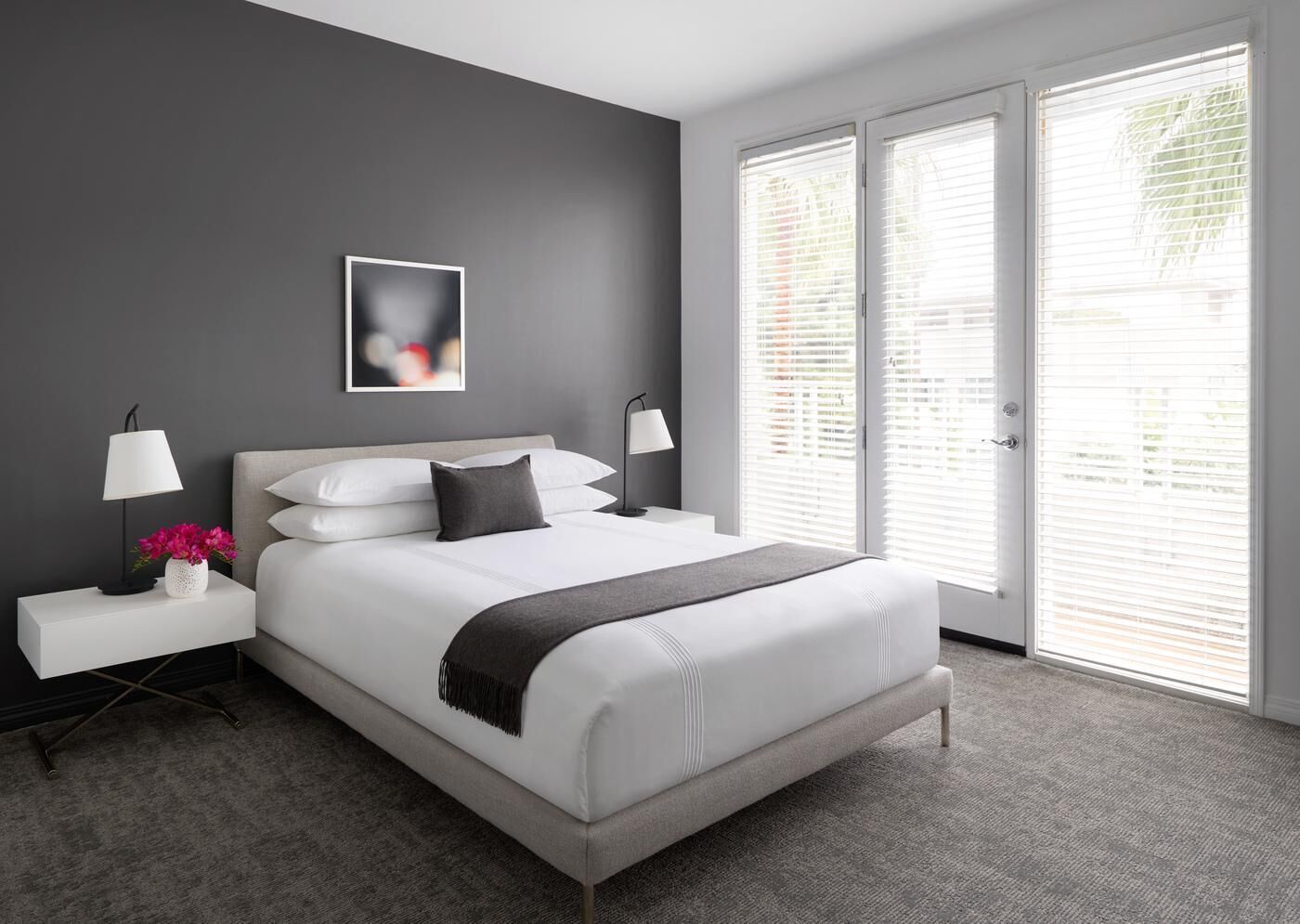 Un dormitorio moderno con una cama grande, dos mesitas de noche, una mesita auxiliar, un cuadro enmarcado y vistas al exterior a través de ventanas amplias.