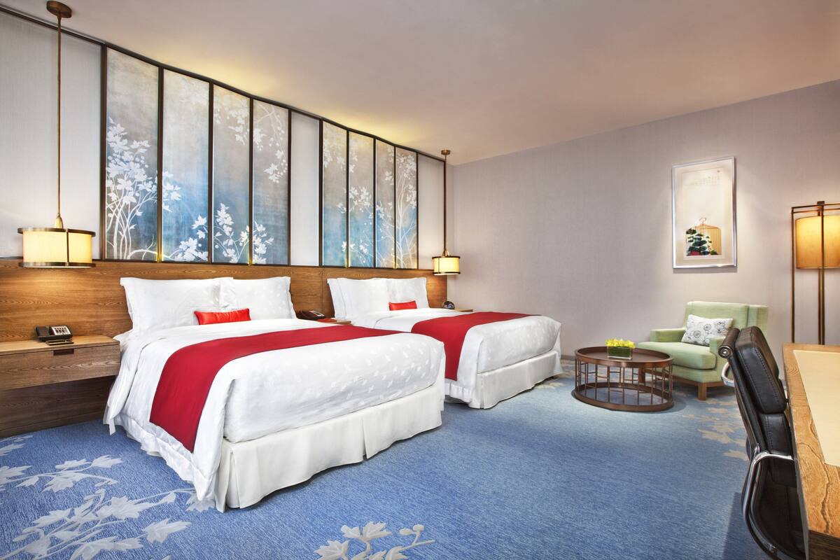 A hotel room with two beds, a desk, a chair, a small table, and a wall-mounted artwork.