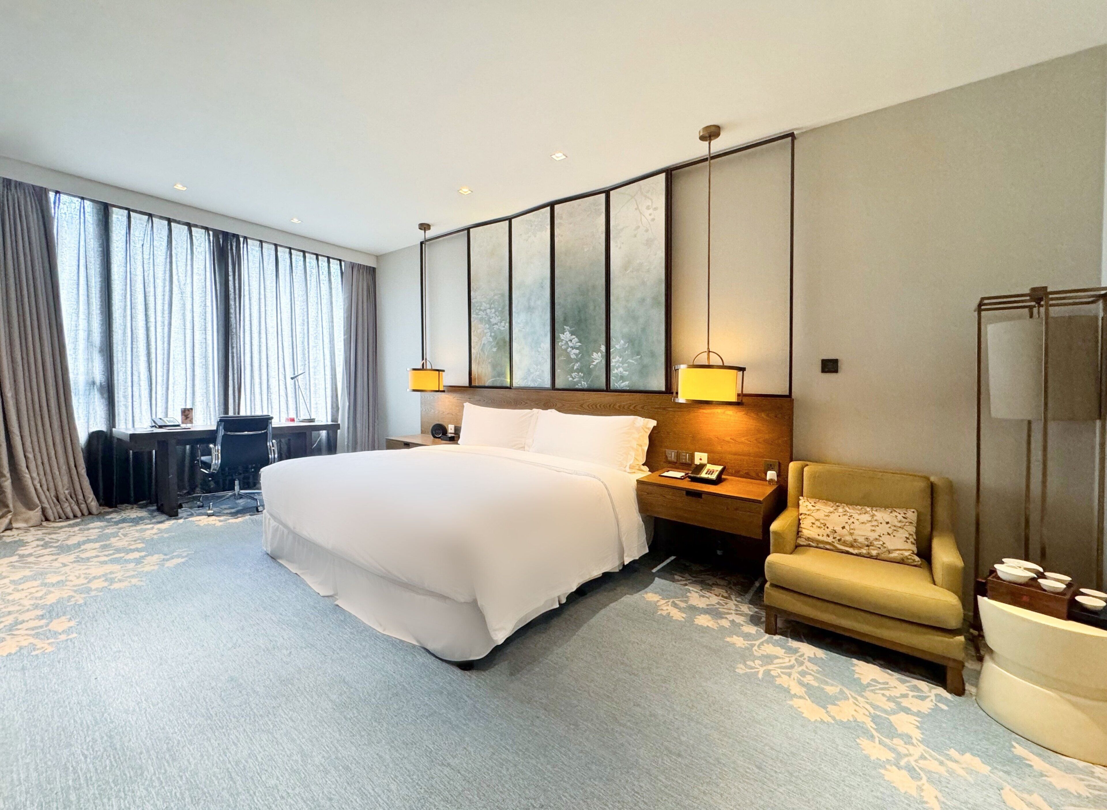 Une chambre d’hôtel avec un grand lit, un bureau, une chaise et une grande fenêtre avec des rideaux.