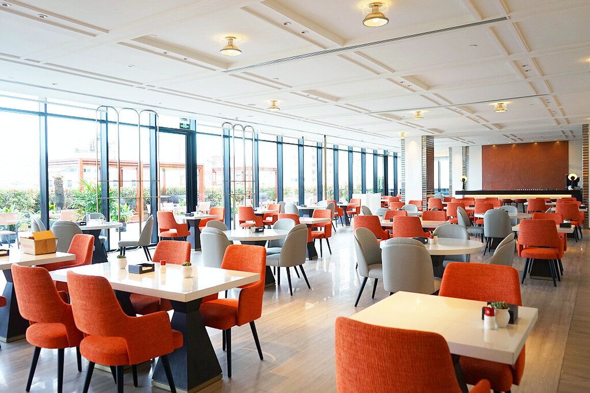 A spacious indoor dining area with multiple tables and chairs, large windows, and a stage for presentations.