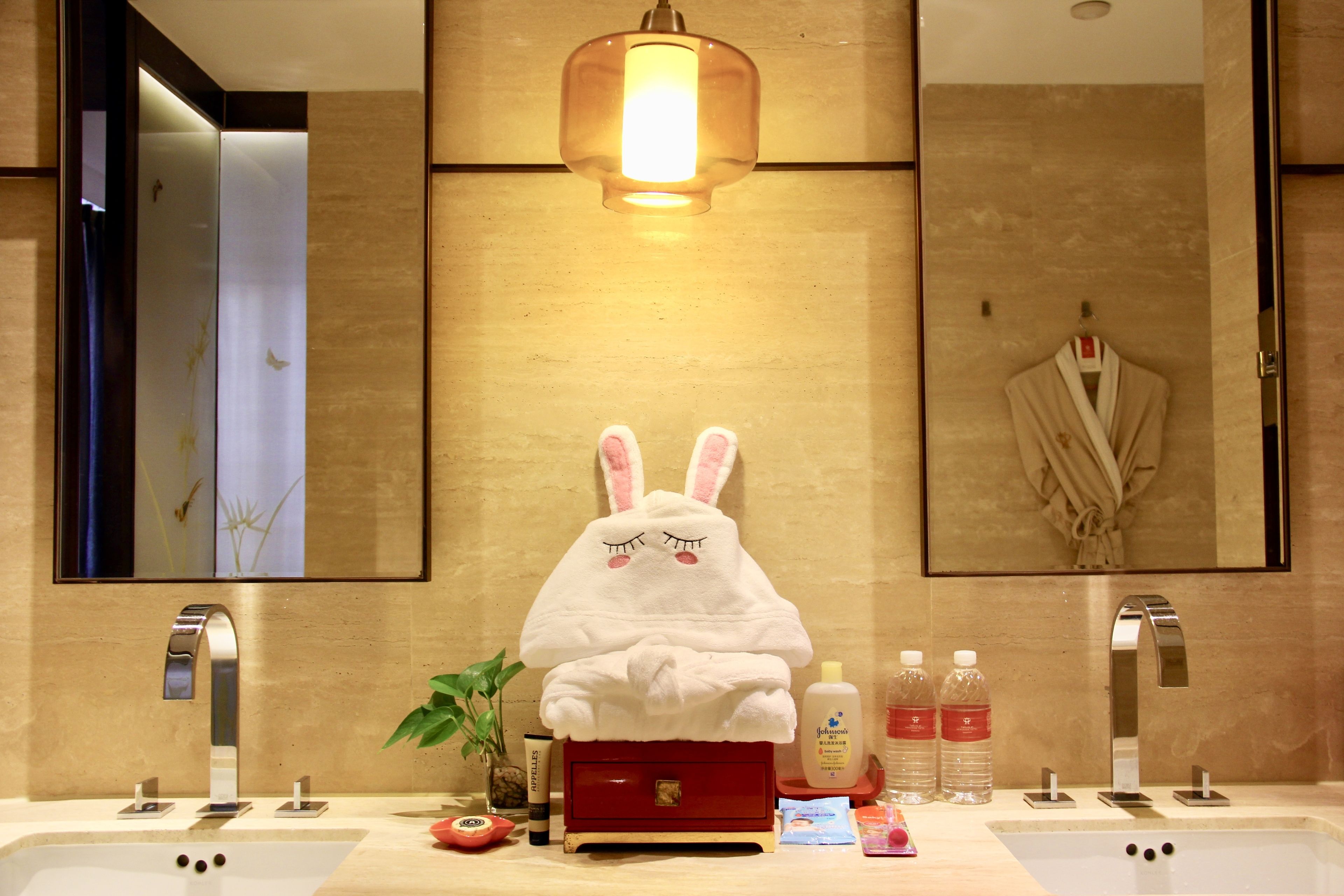 Une salle de bain avec une serviette blanche en forme de lapin posée sur un meuble rouge, un miroir, deux lavabos et des articles de toilette sur le comptoir.