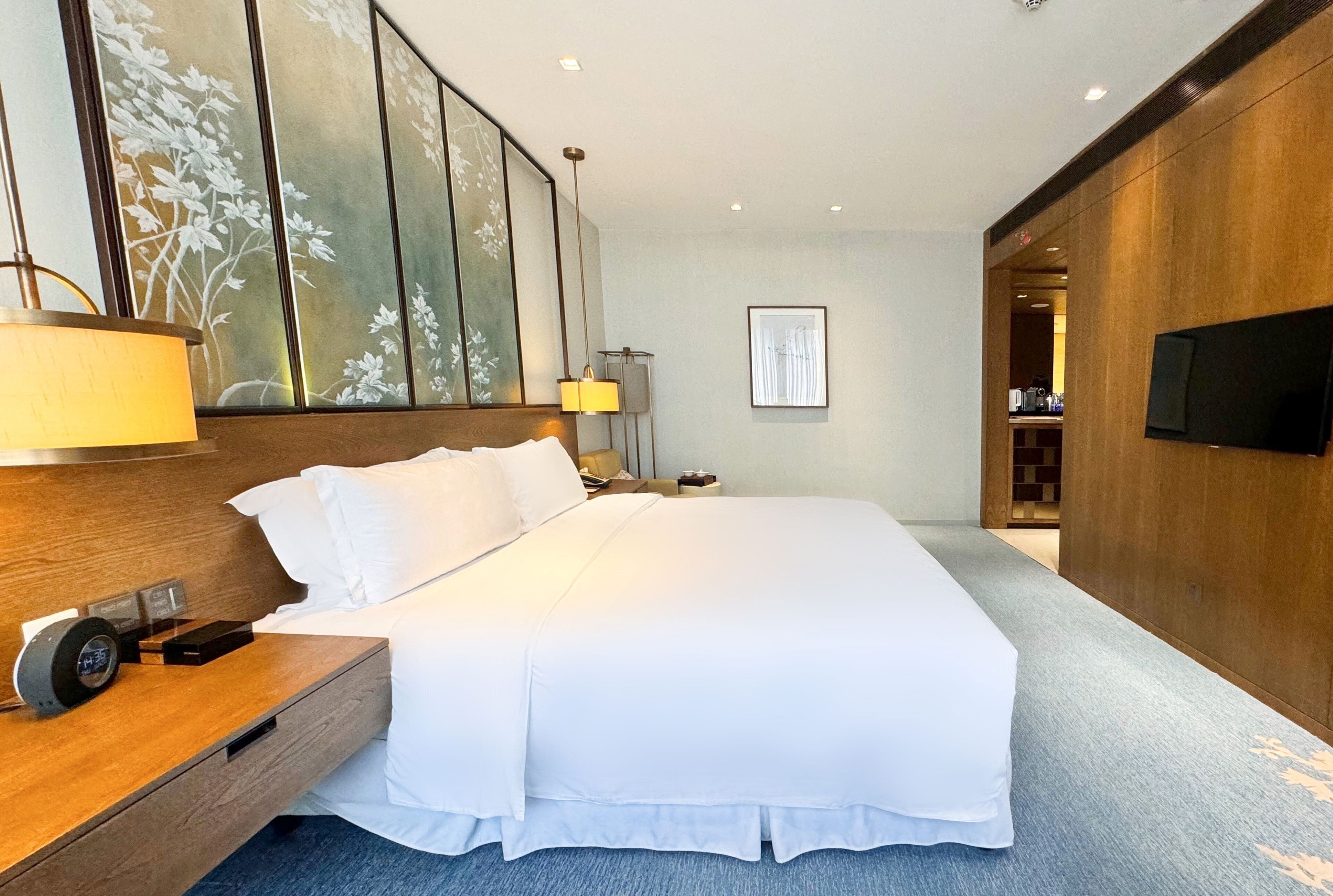 Quarto de hotel com uma cama grande, mesa de cabeceira, abajur e televisão na parede.