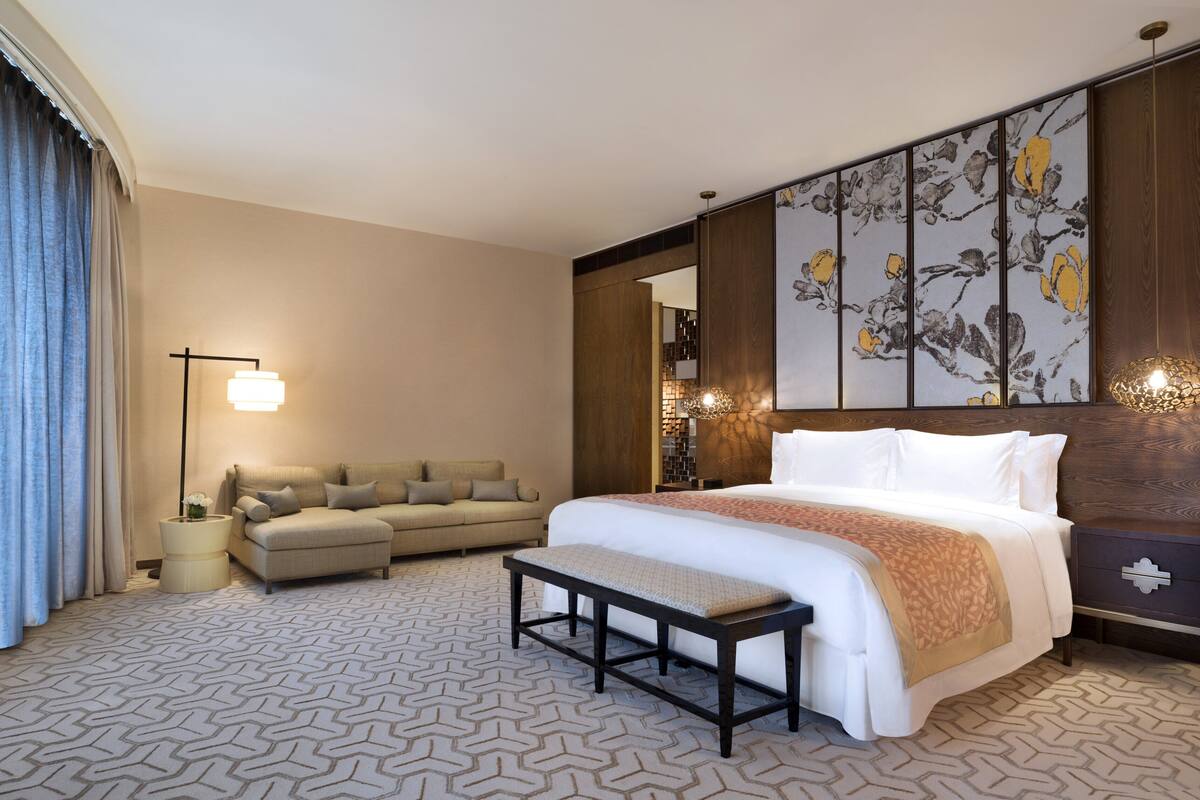 A hotel room with a large bed, a sofa, a bench, a nightstand, and a wall with a floral design.