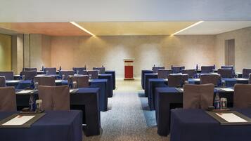 Un salón de conferencias con filas de mesas y sillas, un atril y botellas de agua sobre cada mesa.