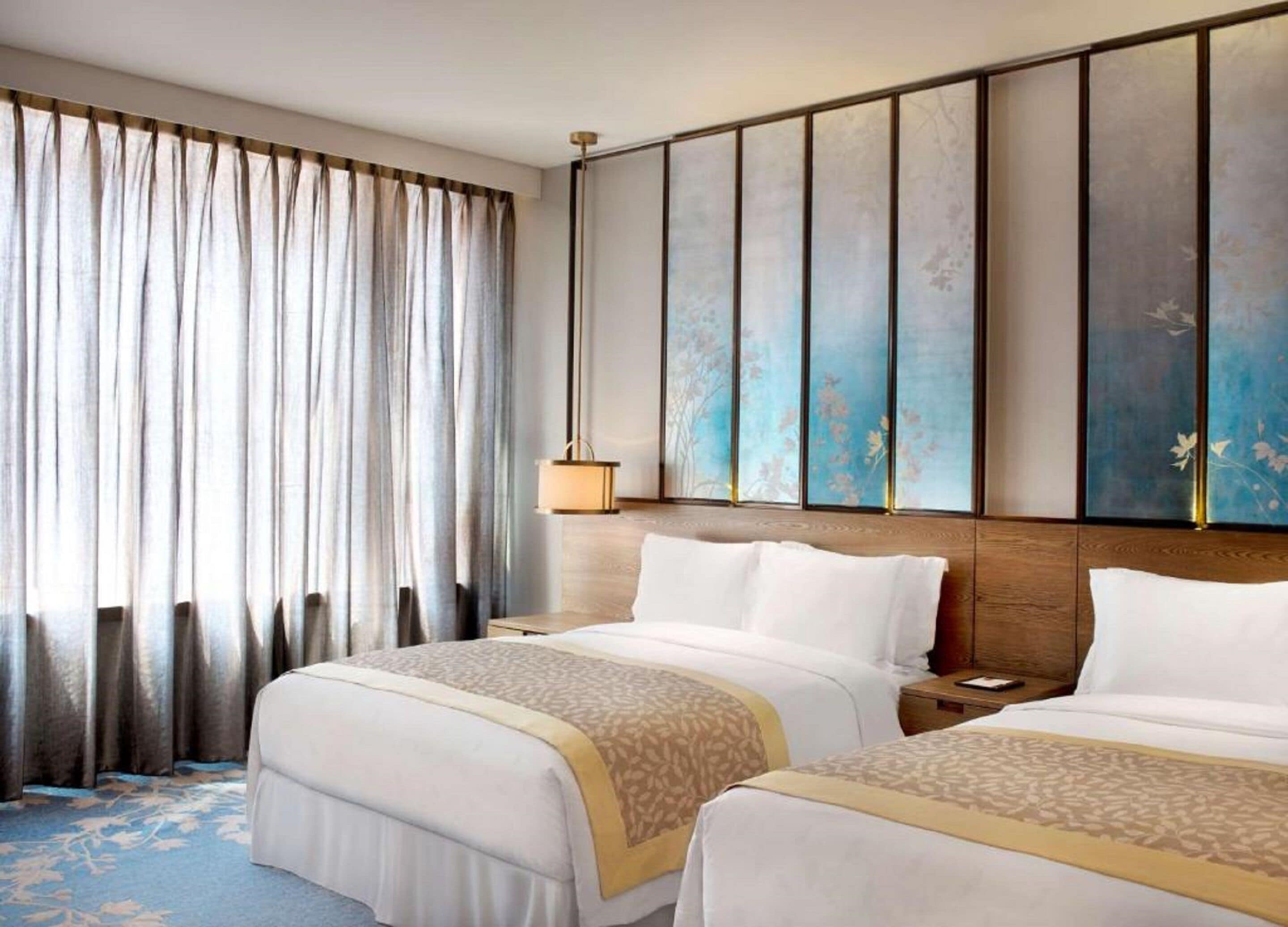 A hotel room with two beds, a large window with curtains, a wall-mounted artwork, and a bedside table with a lamp.