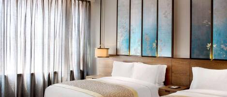 A hotel room with two beds, a large window with curtains, a wall-mounted artwork, and a bedside table with a lamp.