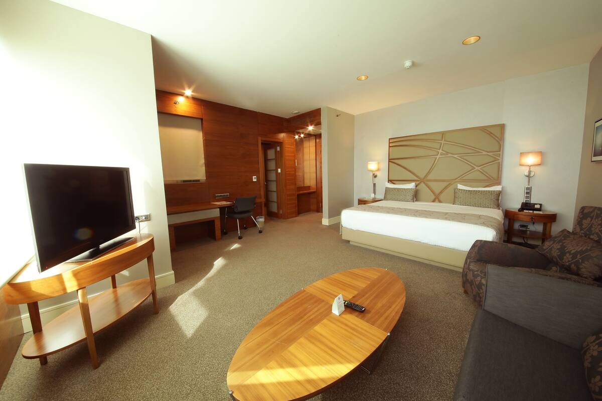 A hotel room with a TV on a wooden stand, a bed with a headboard, a sofa, a desk with a chair, and a small table with a lamp.