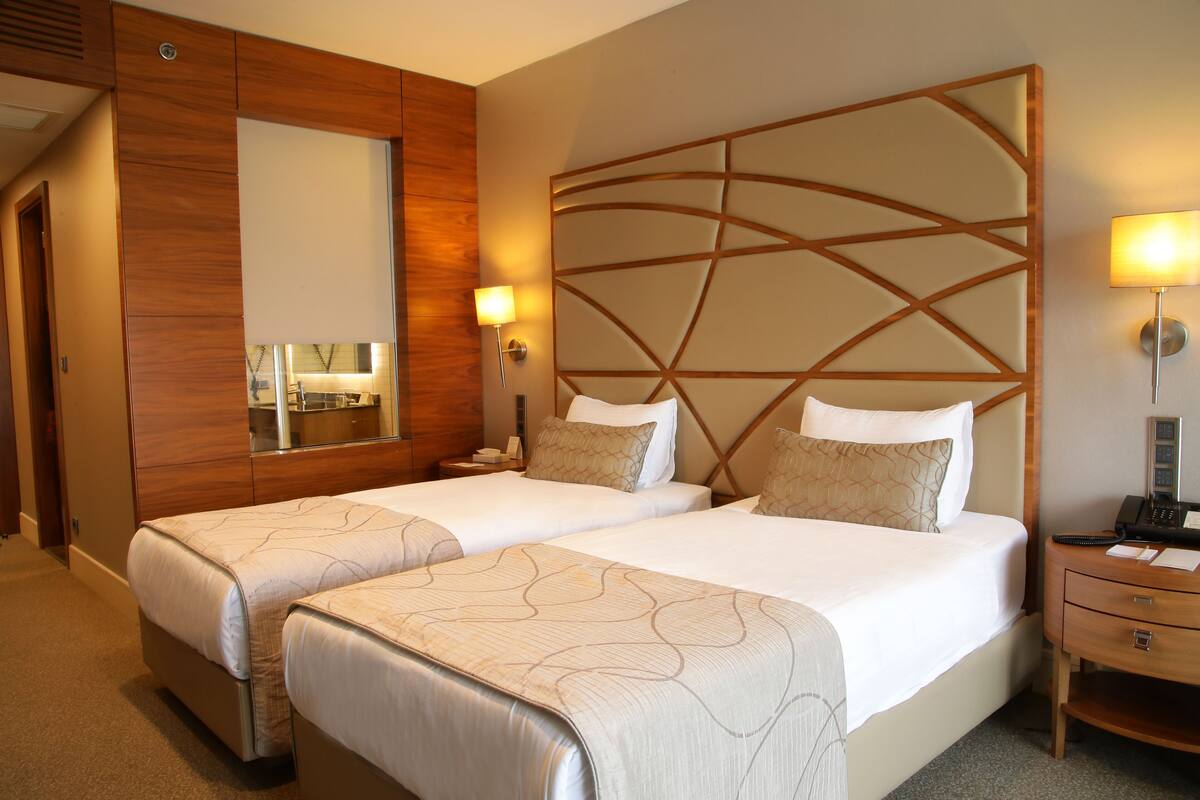 A hotel room with two beds, a wooden headboard, bedside lamps, a mirror, and a desk with a phone.