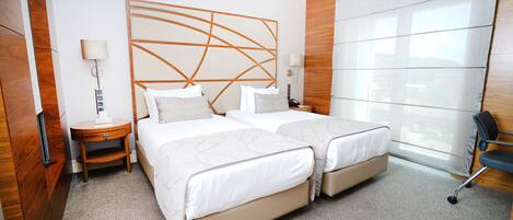 A hotel room with two beds, a wooden headboard, a desk with a lamp, and a large window.