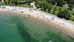 An aerial view of a beach with people swimming and sunbathing, a road adjacent to the beach, and greenery in the background.