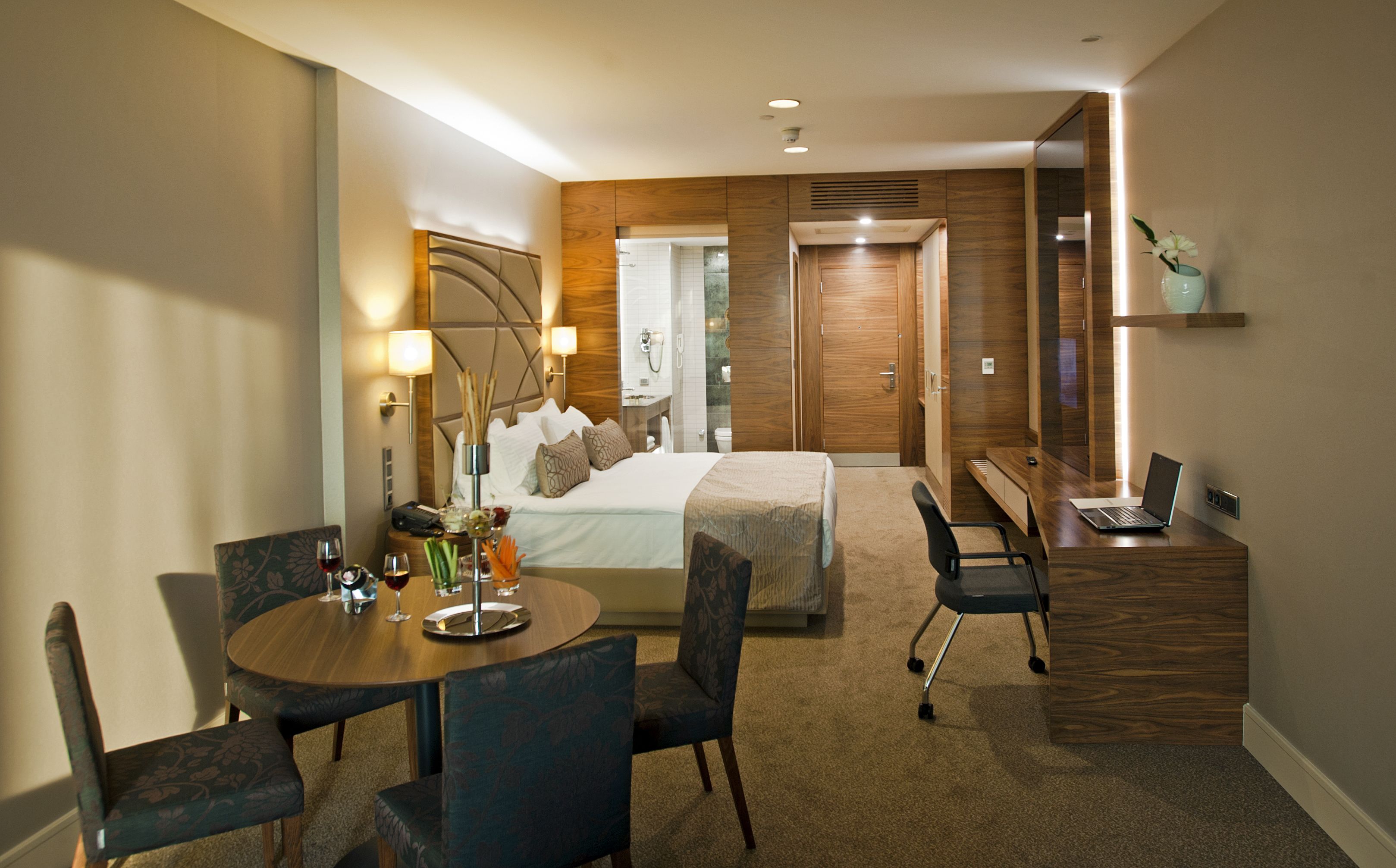A hotel room with a bed, desk, chairs, and a small table with a vase and candles.
