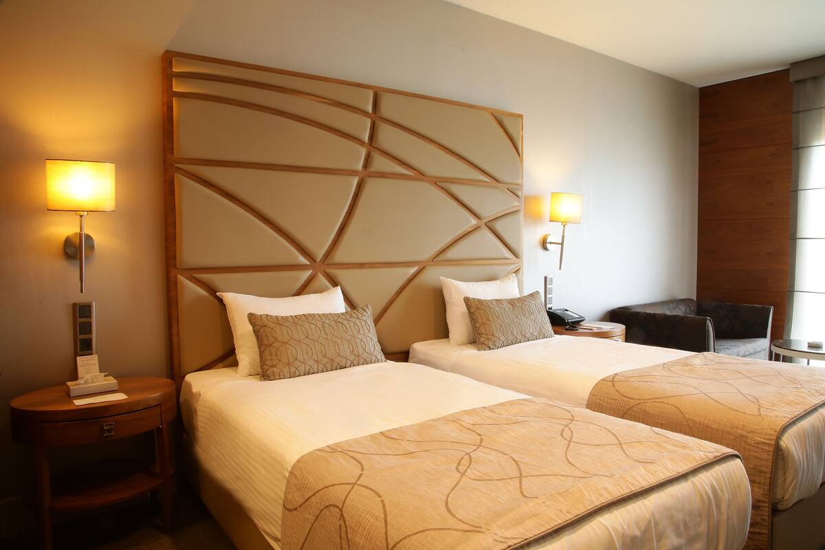 A hotel room with two beds, a wooden headboard, bedside lamps, a desk, and a chair.