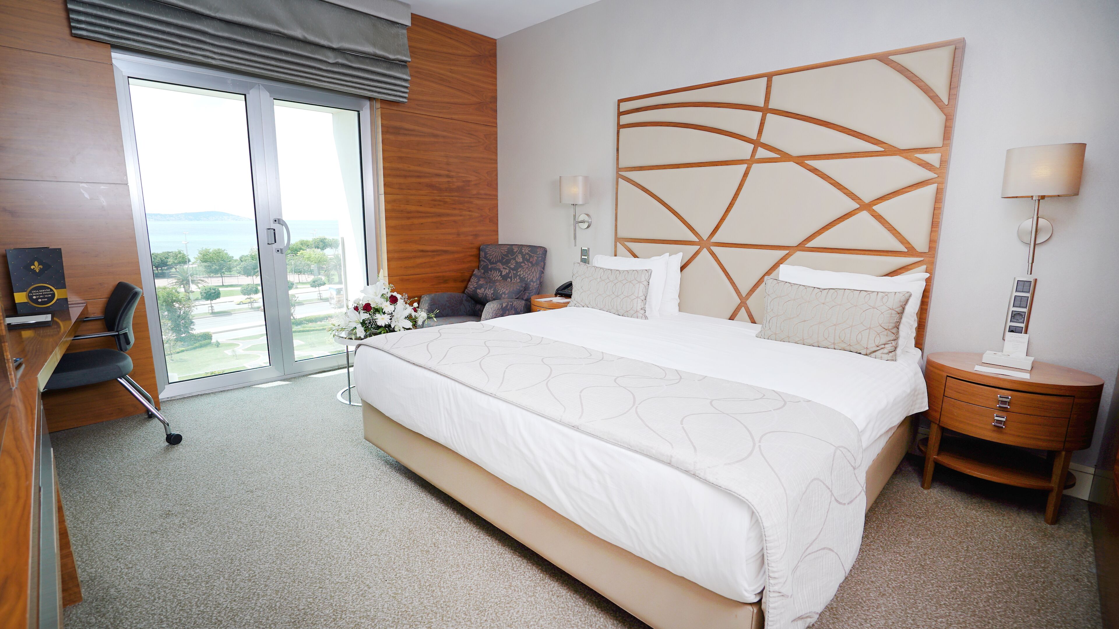 A hotel room with a large bed, a desk with a chair, a nightstand, and a view of the outdoors.