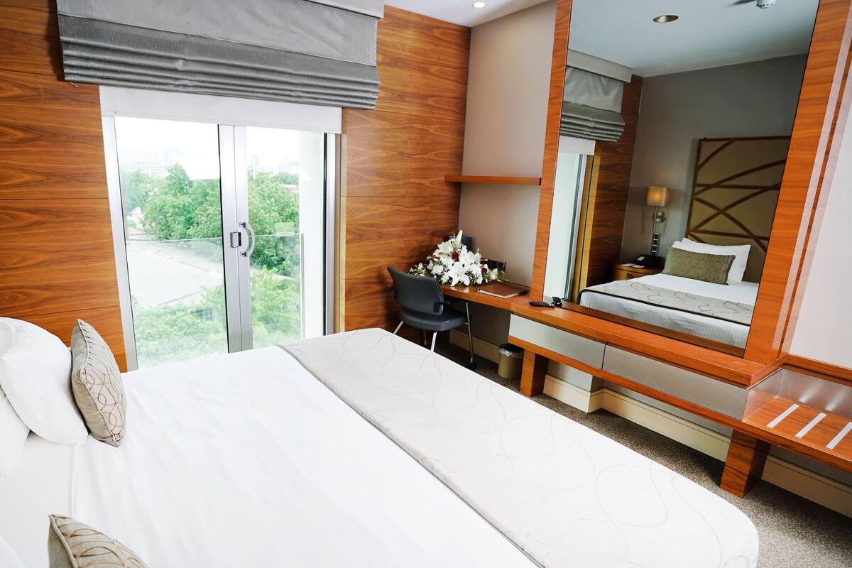A hotel room with a large bed, a desk with a chair, a mirror, and a view of greenery outside.