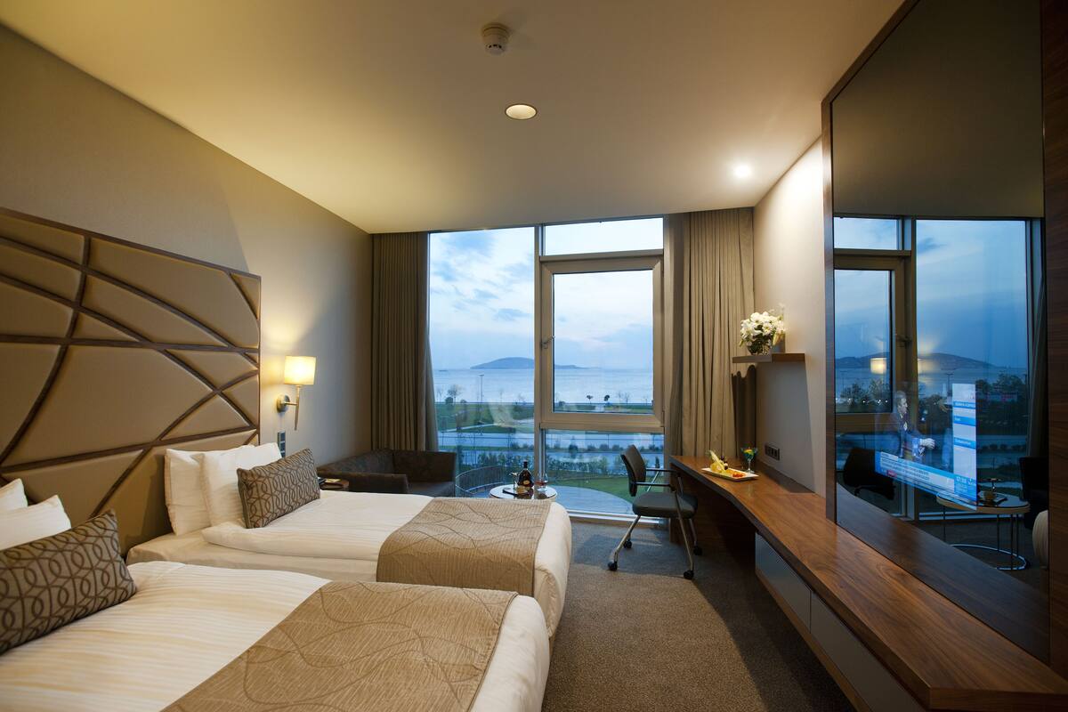 A hotel room with two beds, a desk, a chair, a TV, and a large window offering a view of the outdoors.