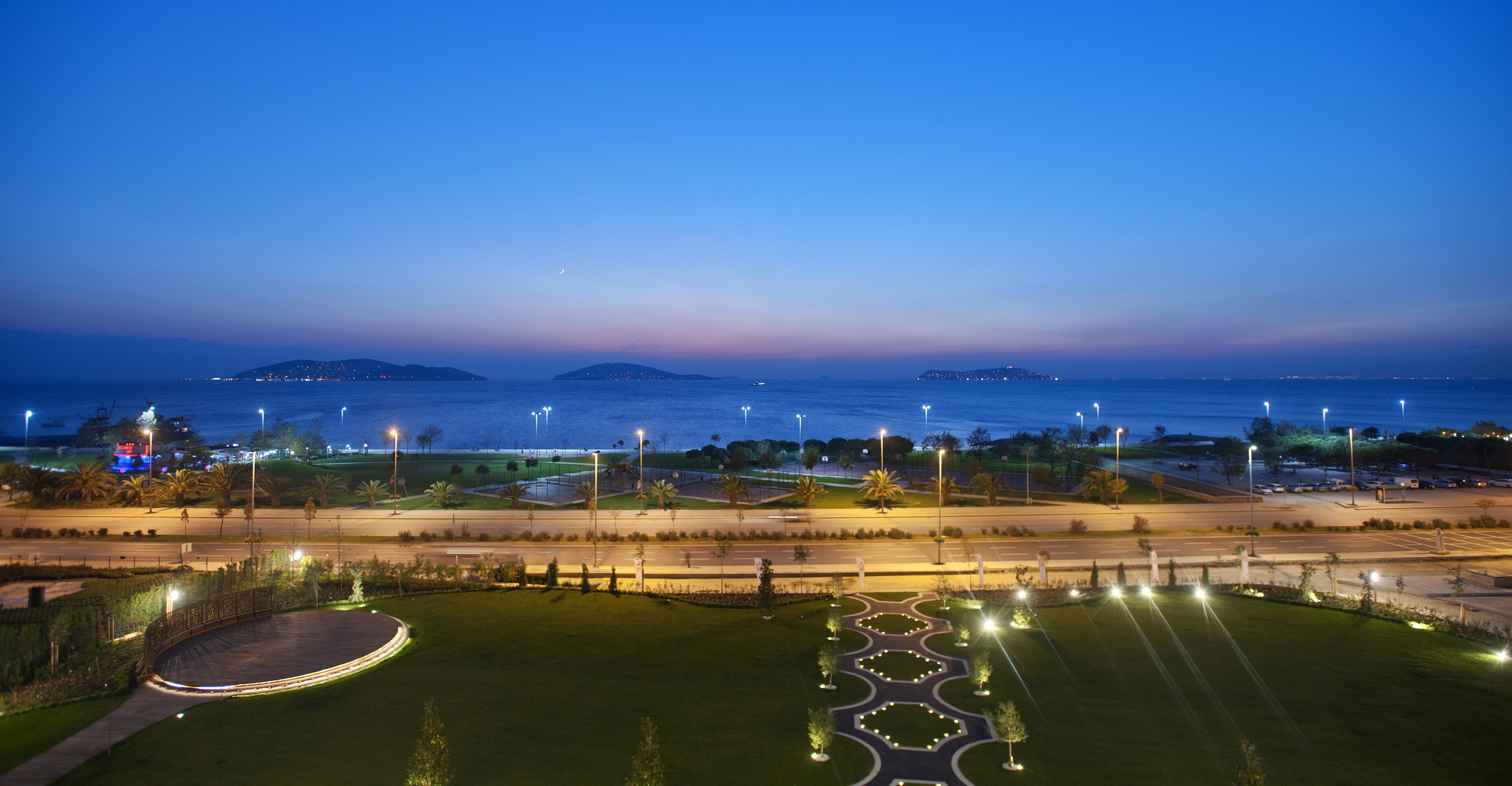 A nighttime view of a coastal area with a road, landscaped gardens, and distant islands.