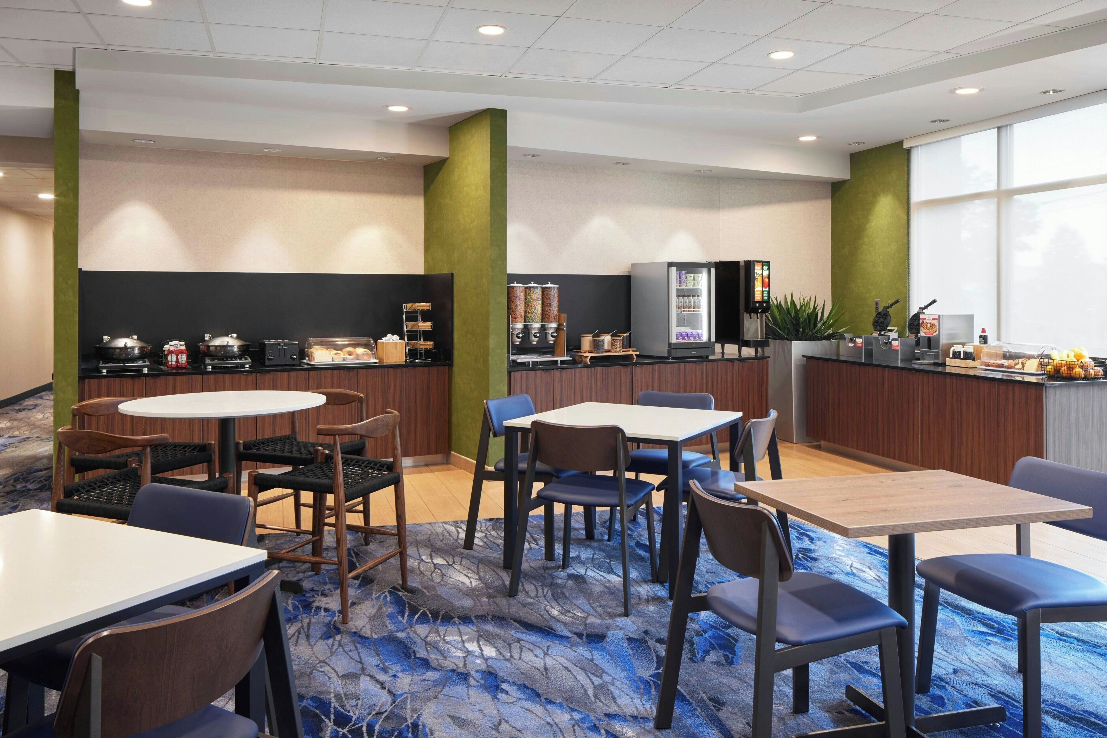 A hotel dining area with round tables, chairs, a refrigerator, and a coffee station.