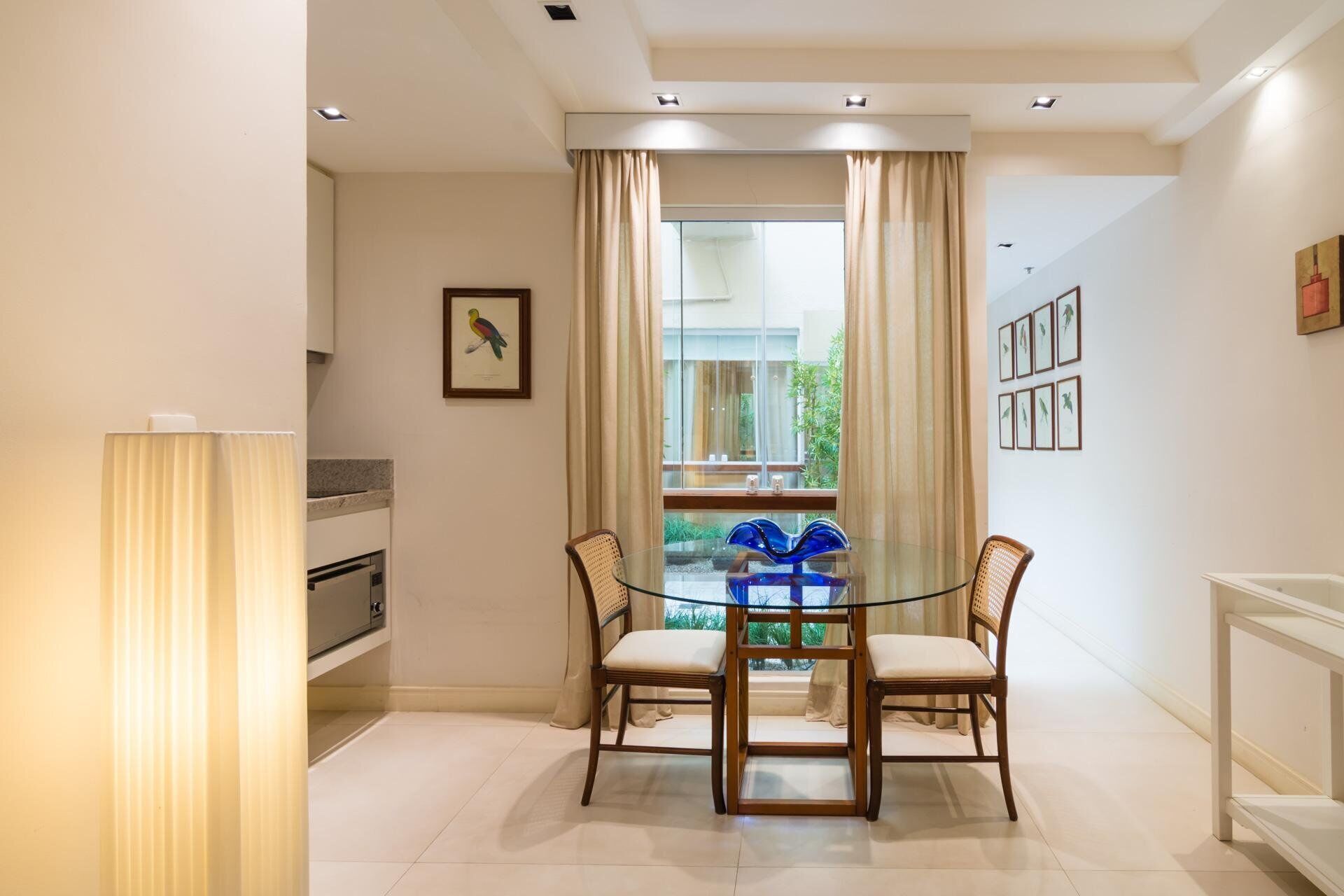 A dining area with a glass table and four chairs, a window with curtains, and framed pictures on the wall.