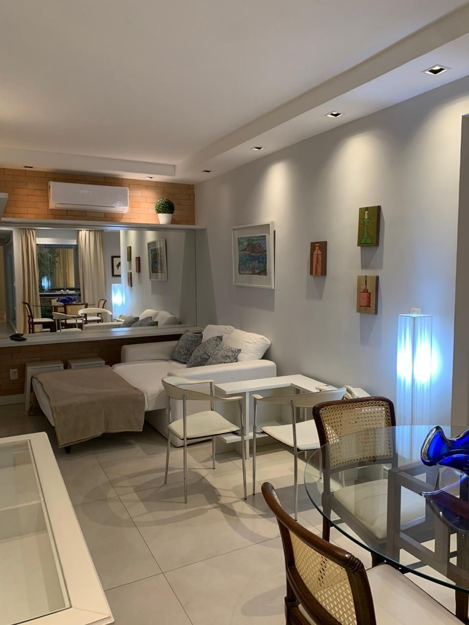 Una sala moderna con un sofá, una mesa de vidrio para comer y una mesa de centro. Hay un ventanal con cortinas, un aire acondicionado instalado en la pared y varias obras de arte enmarcadas.
