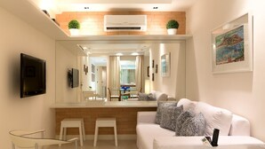 A modern living room with a white sofa, a coffee table, and a TV mounted on the wall. There are two white chairs and a small table with a plant. The room has a large mirror and a painting on the wall. - Mc Flats Ipanema Beach Star (Rio de Janeiro)