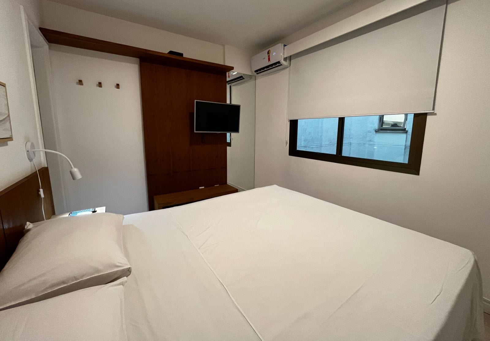 A hotel room with a bed, a television mounted on the wall, a window with a view, and a wooden headboard.