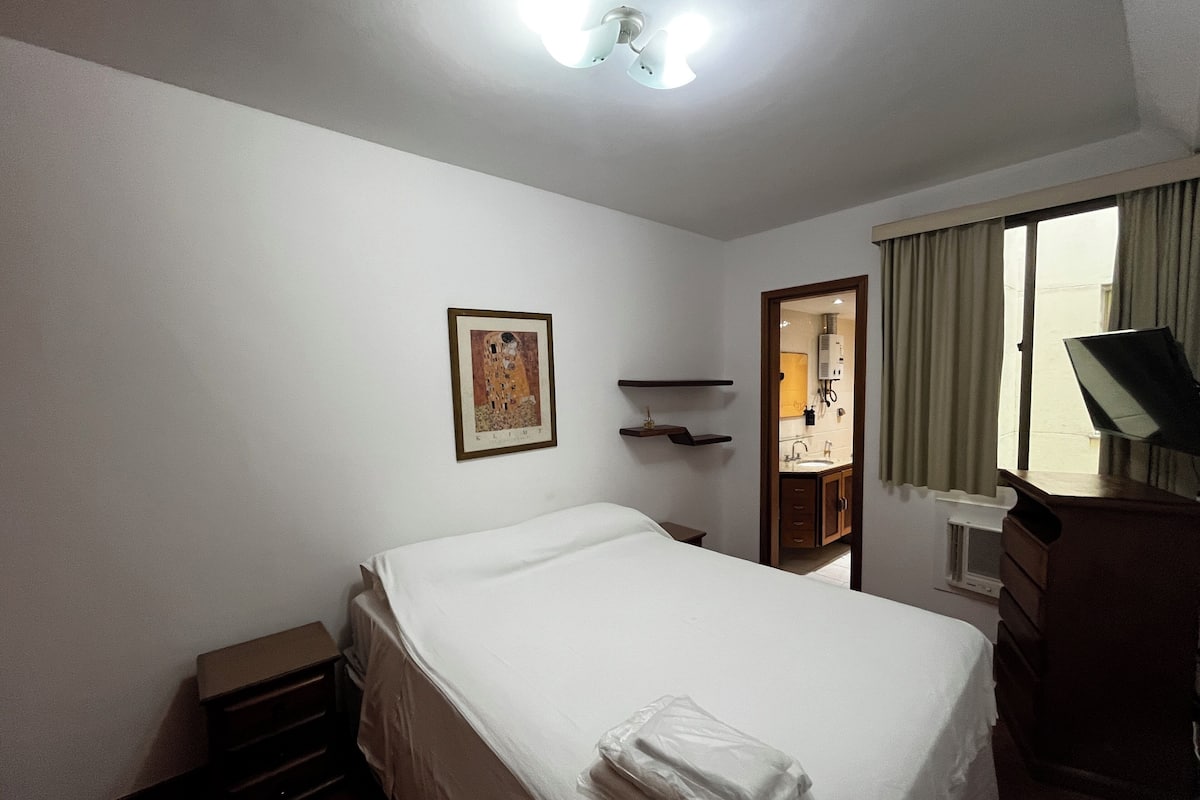 Ein Hotelzimmer mit einem Bett, einem Schreibtisch, einem Stuhl, einem Fenster mit Vorhängen, einem Wandregal und einem gerahmten Bild.