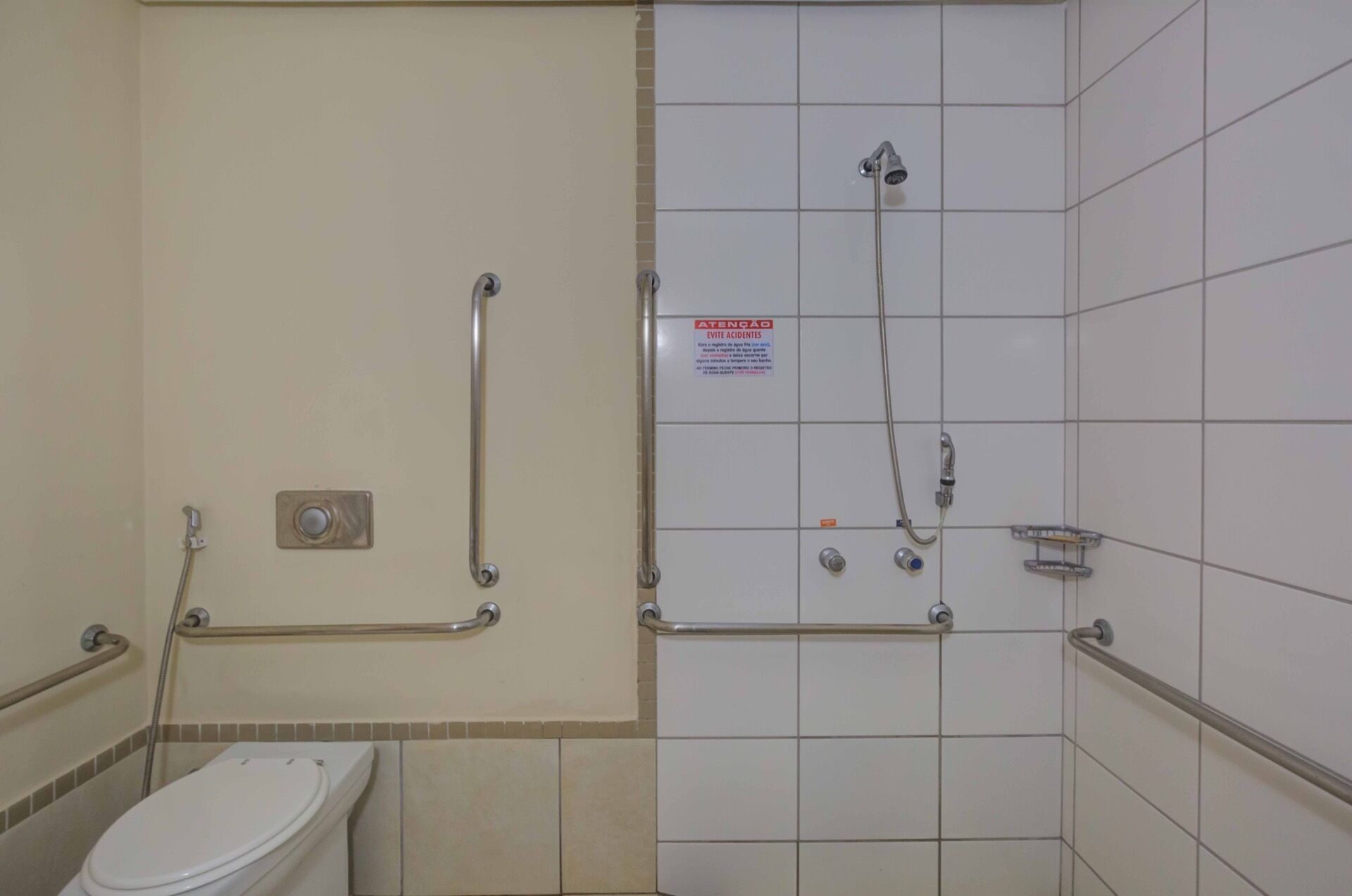 A bathroom with a toilet, grab bars, a shower with a handheld showerhead, and a sign warning of slippery floors.