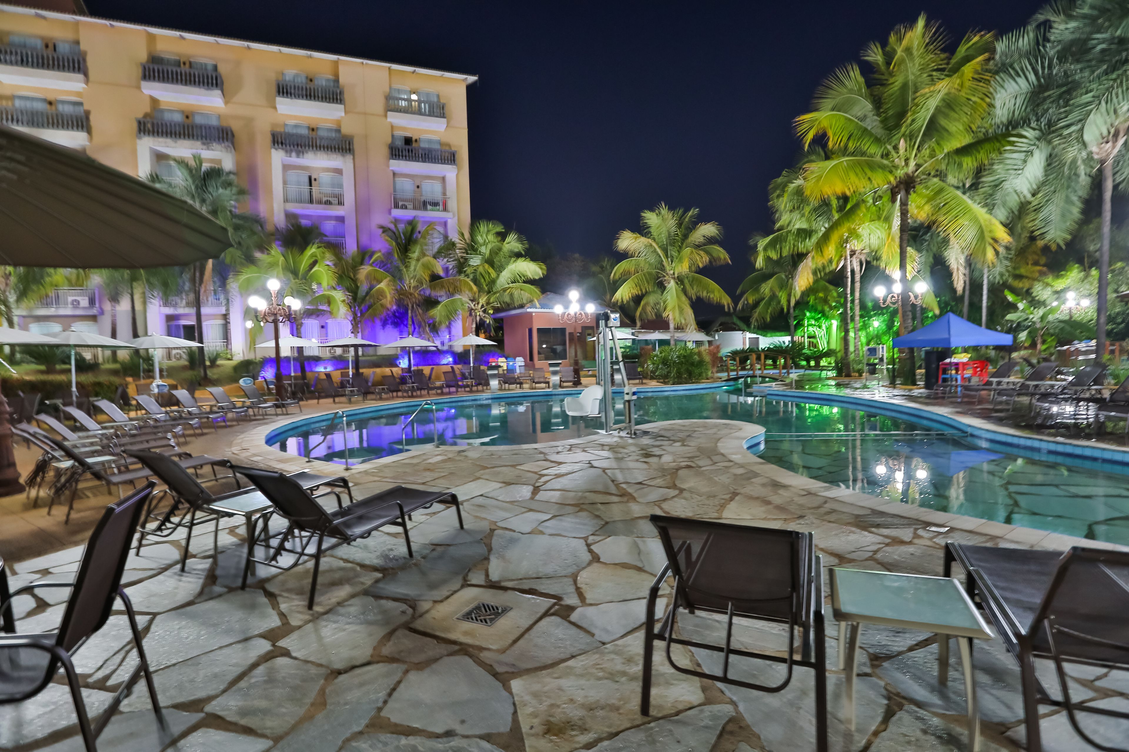 A pool area with lounge chairs and umbrellas, surrounded by palm trees and a building with balconies.