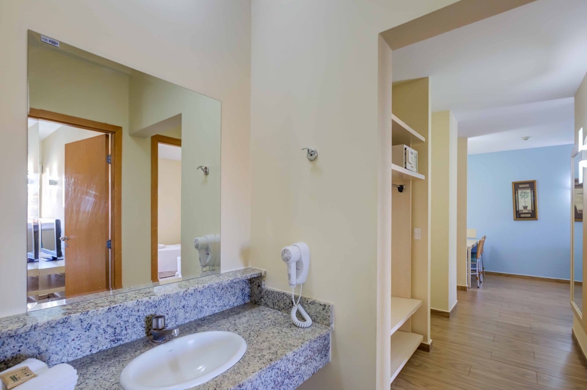 A hotel bathroom with a granite countertop, a white sink, a hairdryer, and a mirror.