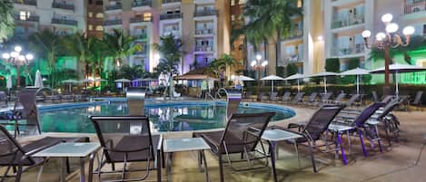 Zona de piscina con sillas de descanso y sombrillas, rodeada de edificios hoteleros de varios pisos con balcones.