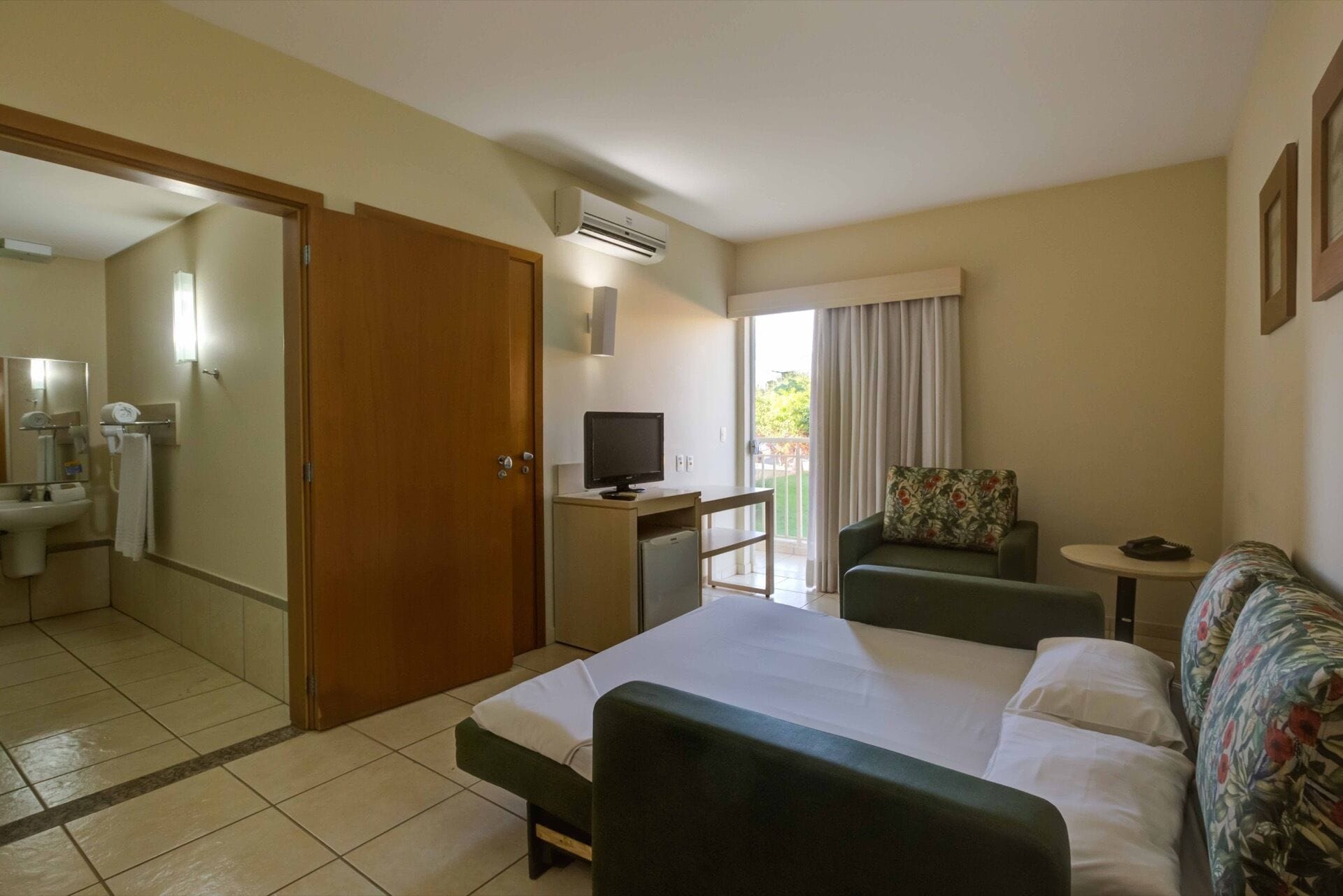 Une chambre d’hôtel comprenant un lit, un bureau avec une télévision, une petite kitchenette, une salle de bain avec un lavabo et un miroir, et un balcon avec vue.