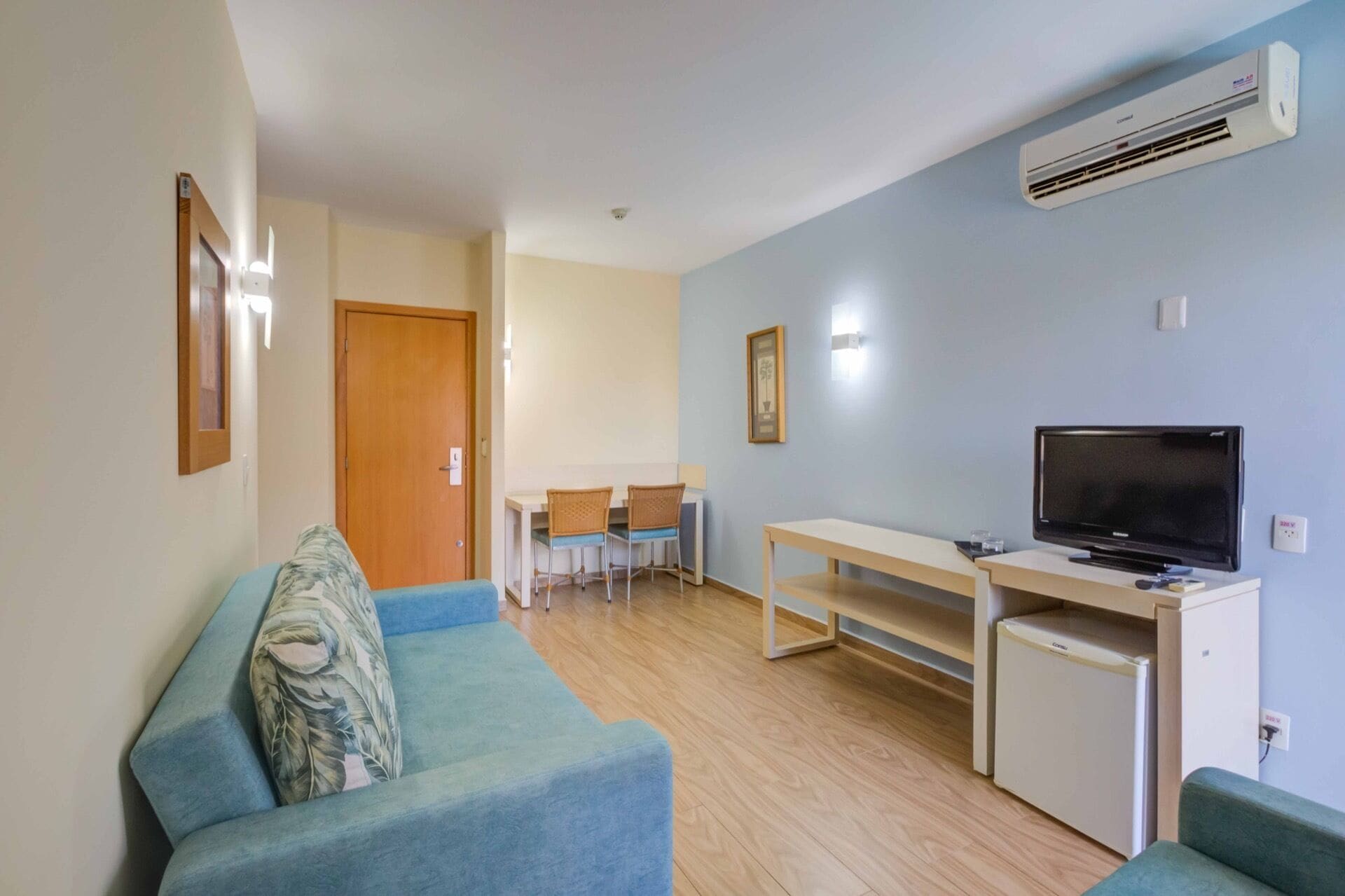 A compact living space with a sofa, a TV, a small fridge, and a dining area with chairs and a table.