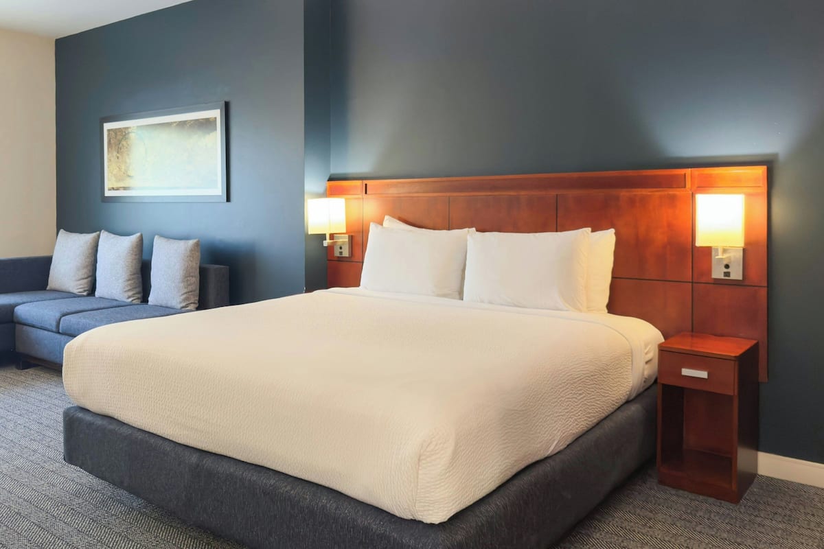 A hotel room with a large bed, a dark-colored sofa, two bedside lamps, and a framed picture on the wall.