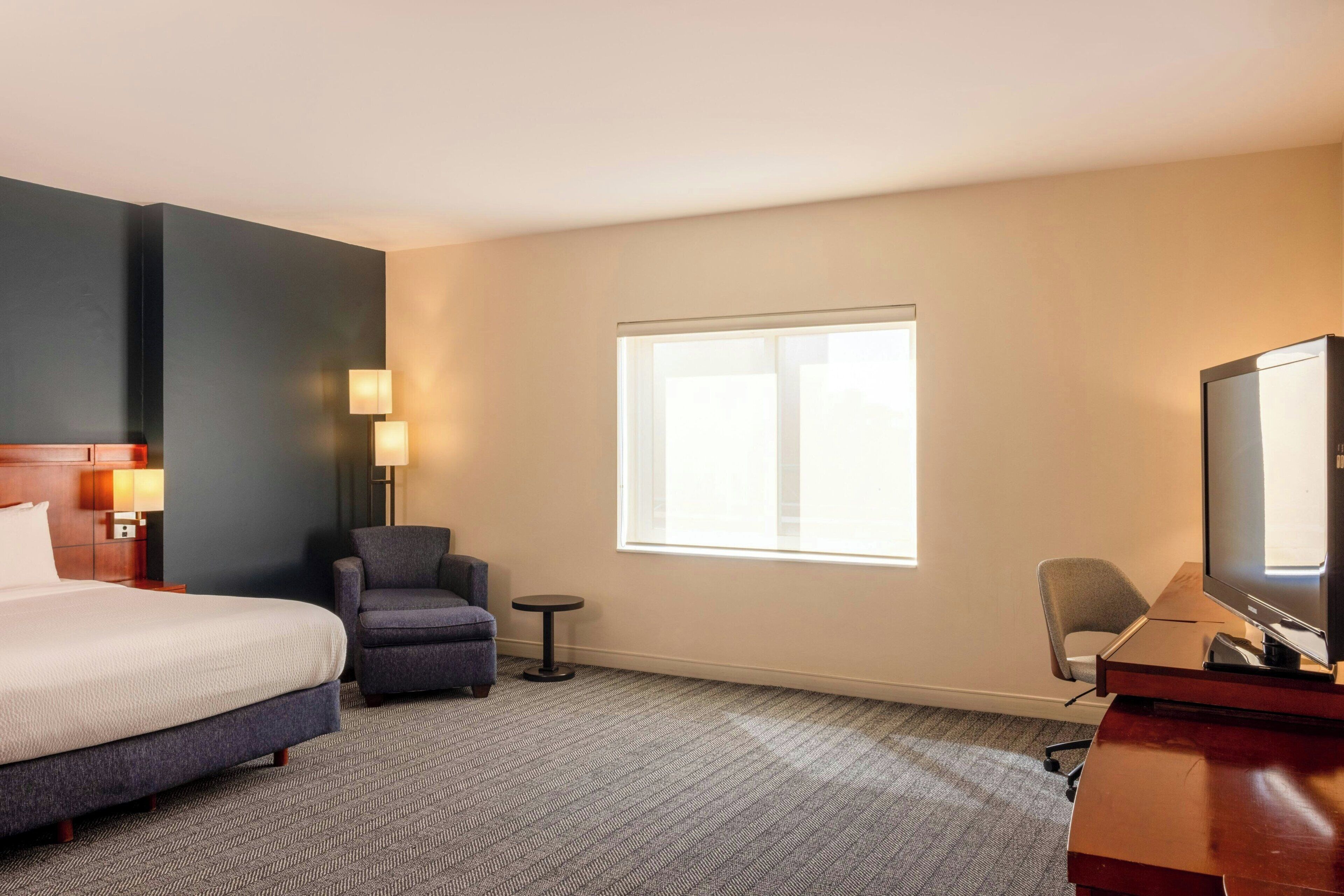 Een hotelkamer met een bed, twee fauteuils, een bureau met een televisie en een raam dat natuurlijk licht biedt.