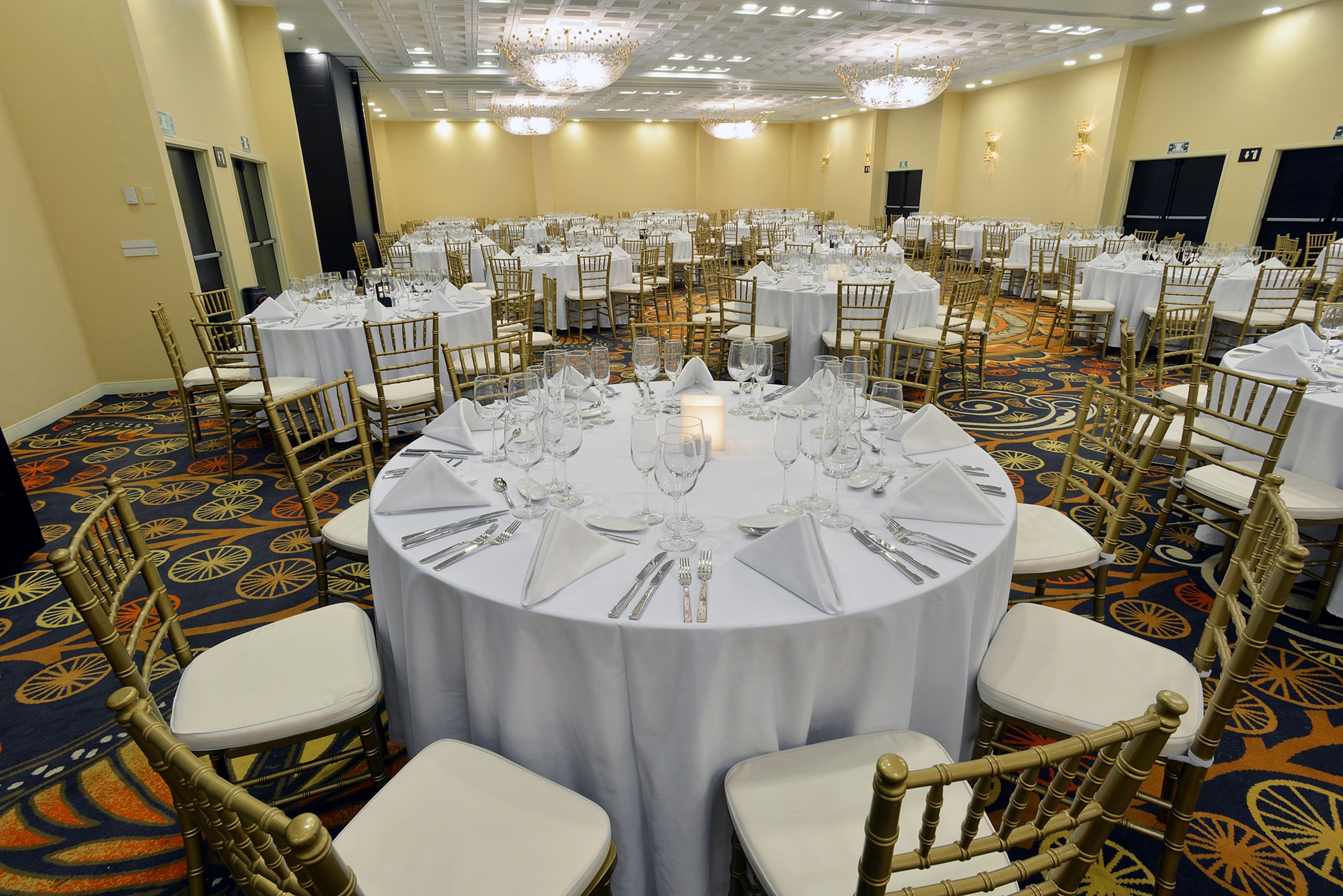 Une salle de banquet avec des tables rondes dressées pour le repas, chacune étant recouverte d’une nappe blanche et entourée de chaises dorées.