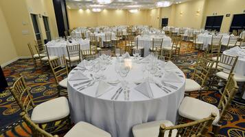 Une salle de banquet avec des tables rondes dressées pour le repas, chacune étant recouverte d’une nappe blanche et entourée de chaises dorées.