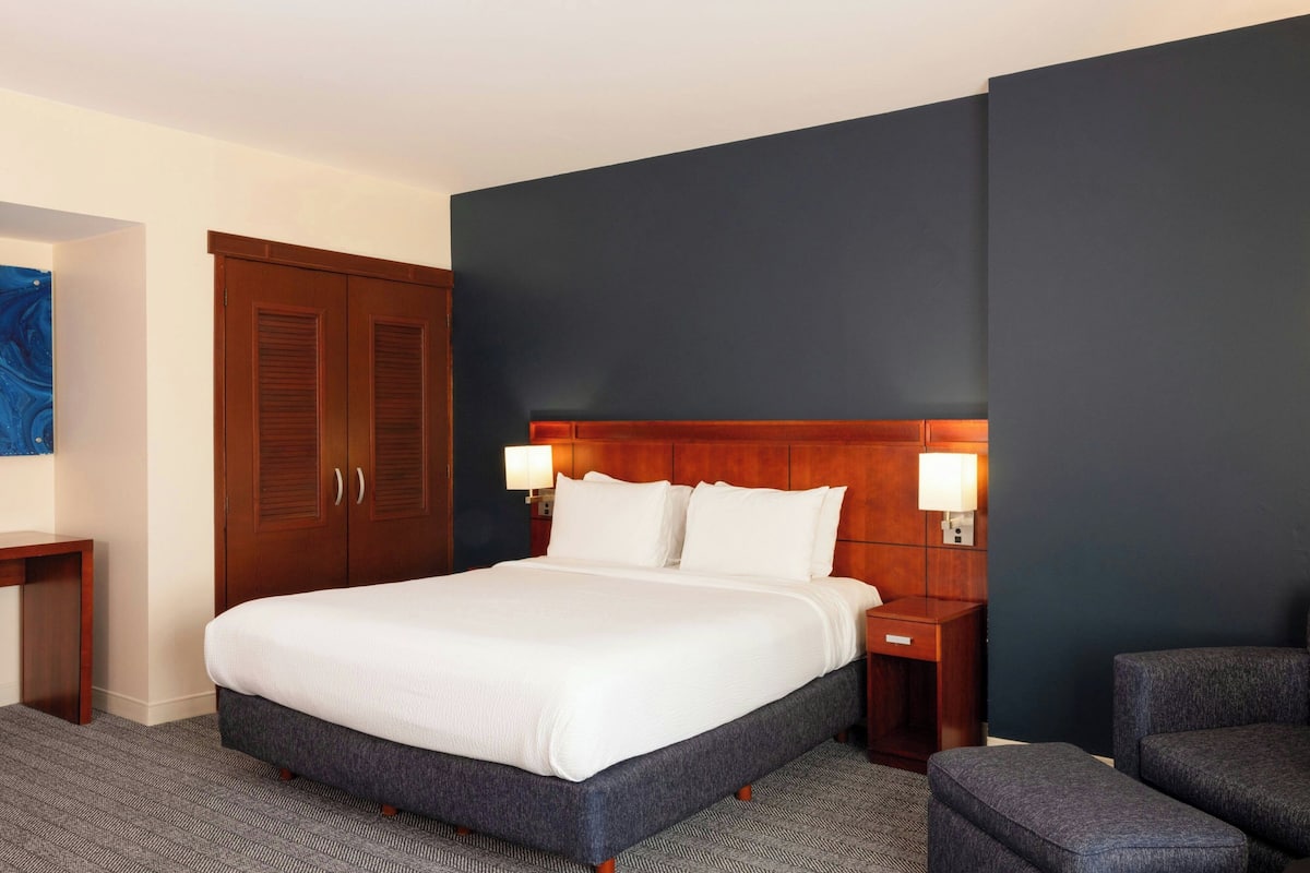 A hotel room with a large bed, wooden headboard, two bedside tables with lamps, a desk, and a chair.