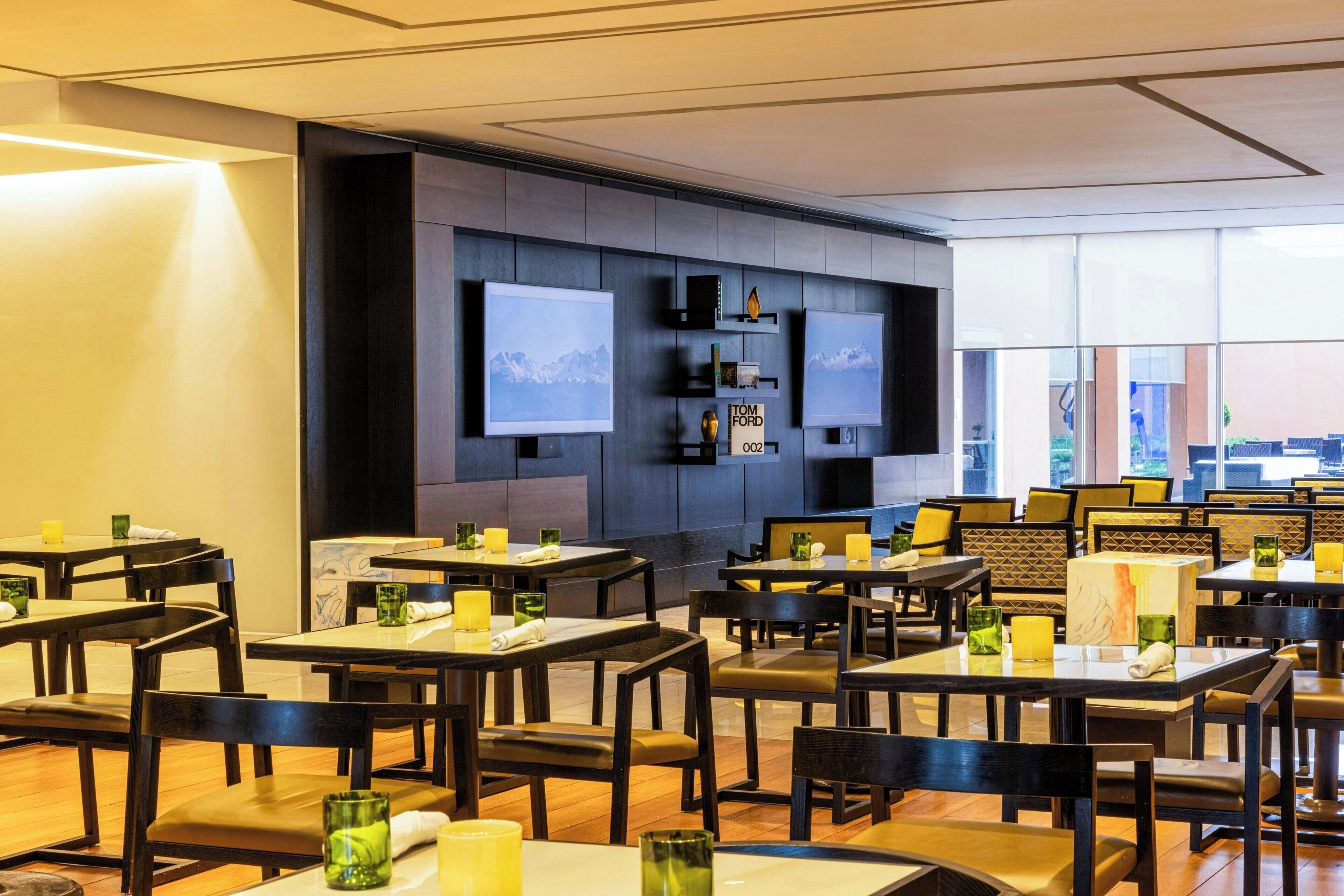 A modern restaurant with multiple tables and chairs, a wall-mounted TV, and a view of the outdoors.