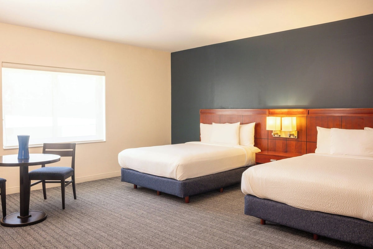 A hotel room with two beds, a small table with a chair, a window, and a nightstand with a lamp.