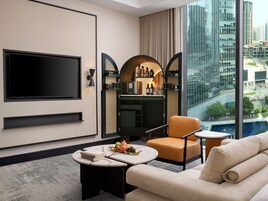 A modern hotel room with a large TV, a bar area, a sofa, and a view of the cityscape.