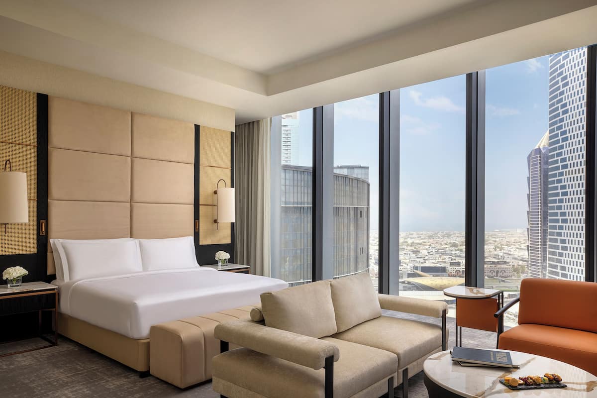 Habitación de hotel moderna con una cama grande, un sofá, una mesita de centro y vistas de la ciudad a través de amplios ventanales.