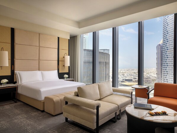 A modern hotel room with a large bed, a sofa, a coffee table, and a view of the cityscape through large windows.