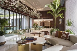 A spacious lobby with a modern design, featuring a mix of seating areas, a large central table, and a variety of plants.