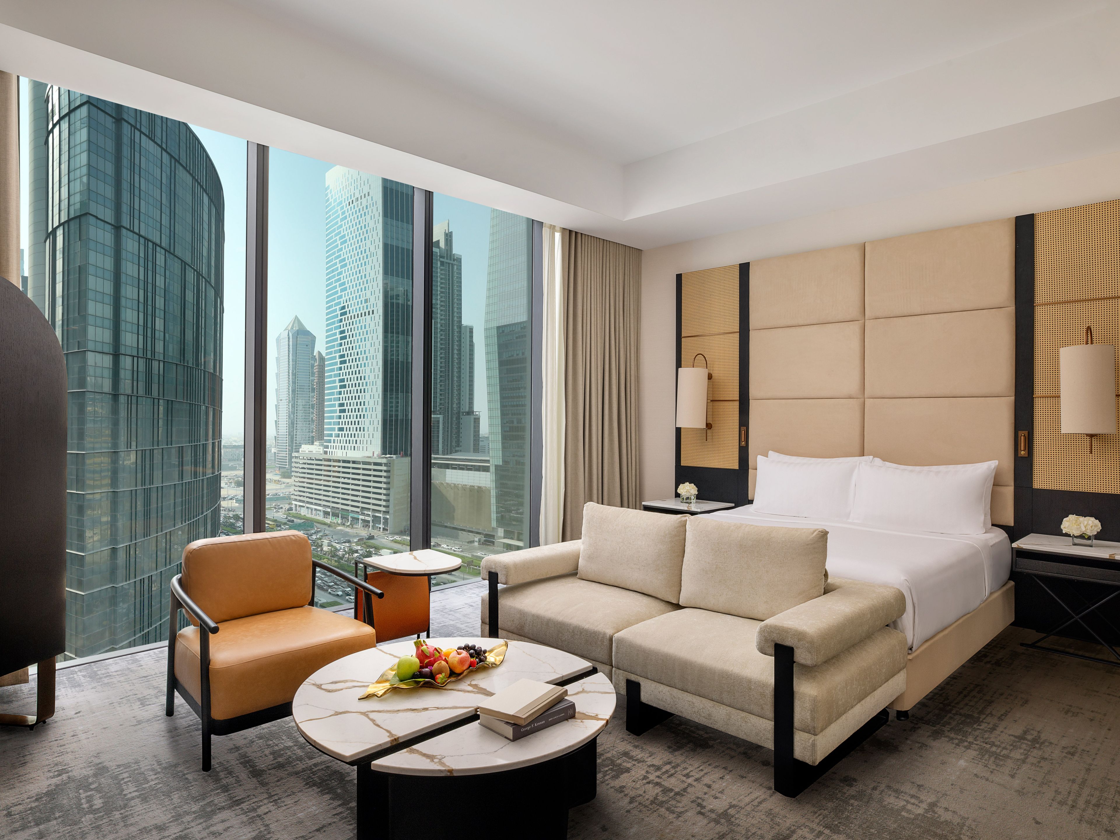 A modern hotel room with a large bed, a sofa, a coffee table, and a view of city skyscrapers.